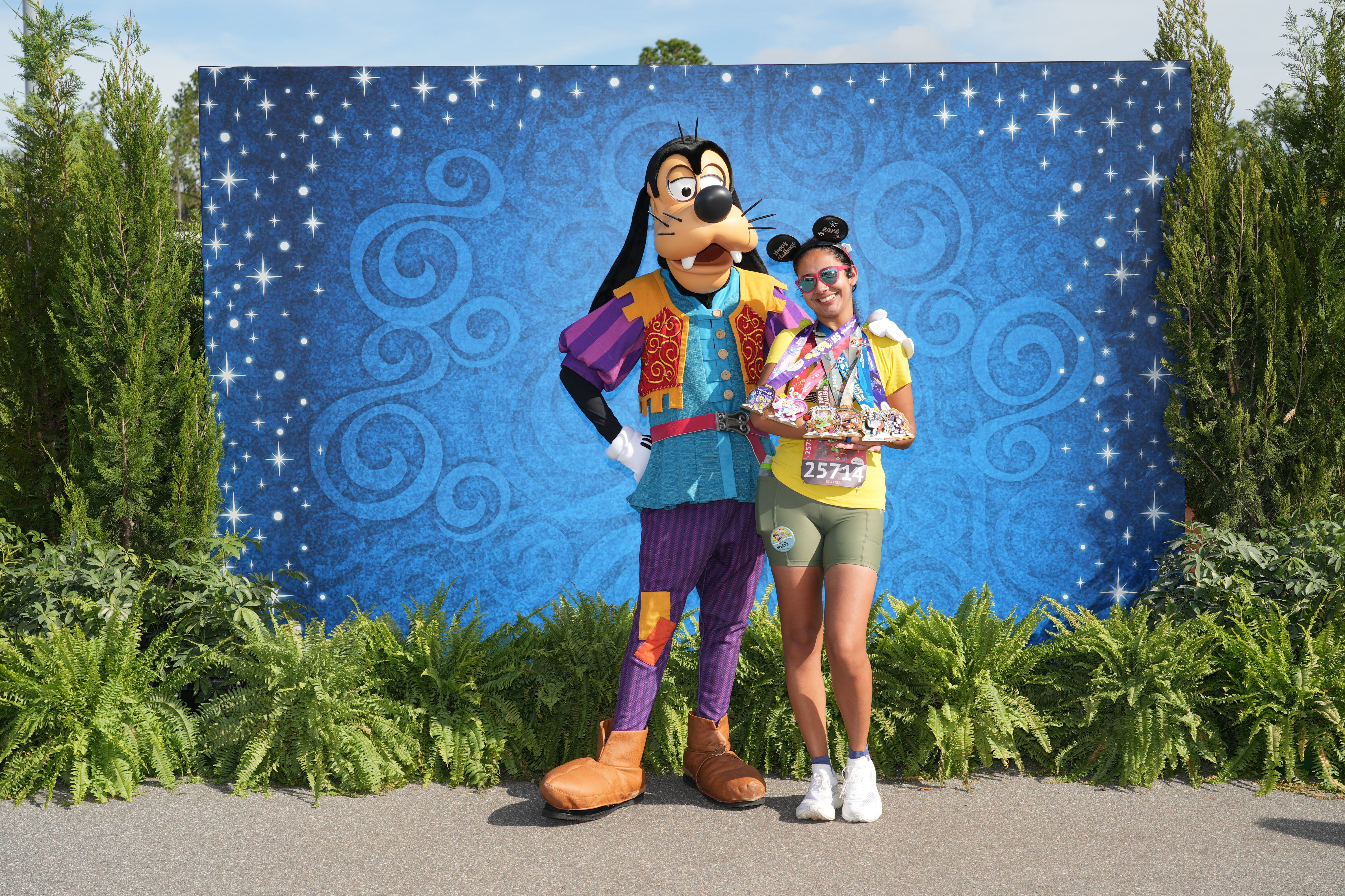 Beatriz da Talla Turismo posa ao lado do Pateta após completar uma corrida da runDisney no Walt Disney World, exibindo suas medalhas e o número de peito da prova