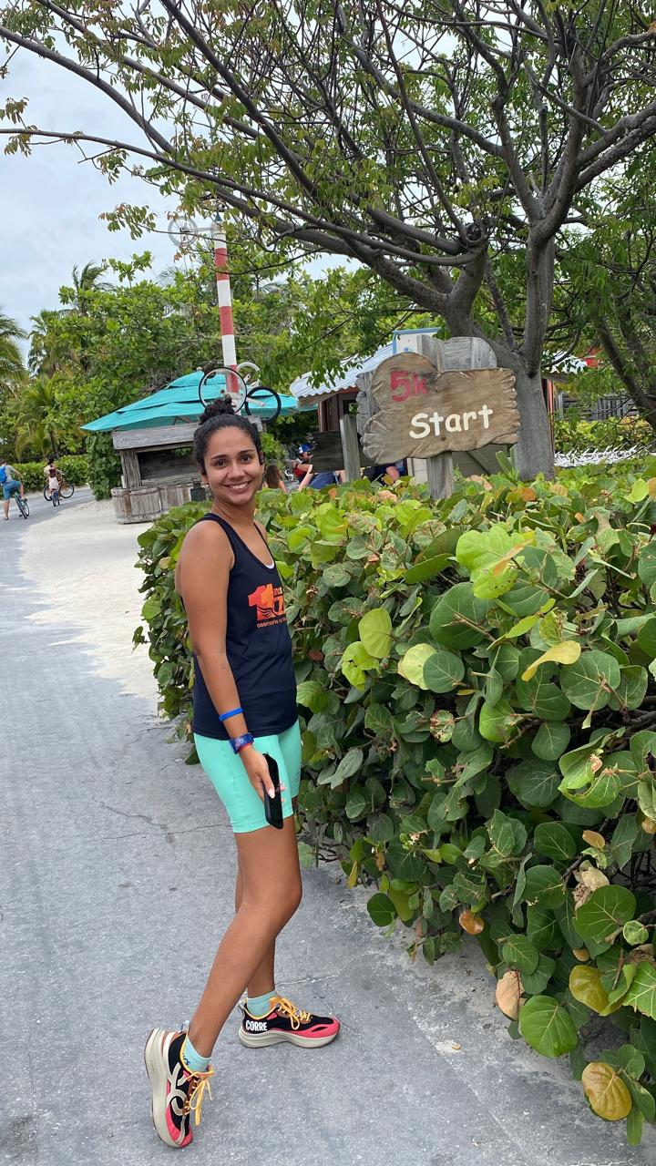 Beatriz da Talla Turismo na ilha Castaway Cay da Disney nas Bahamas, ao lado da placa de largada do percurso 5K, destacando experiências exclusivas do cruzeiro Disney.
