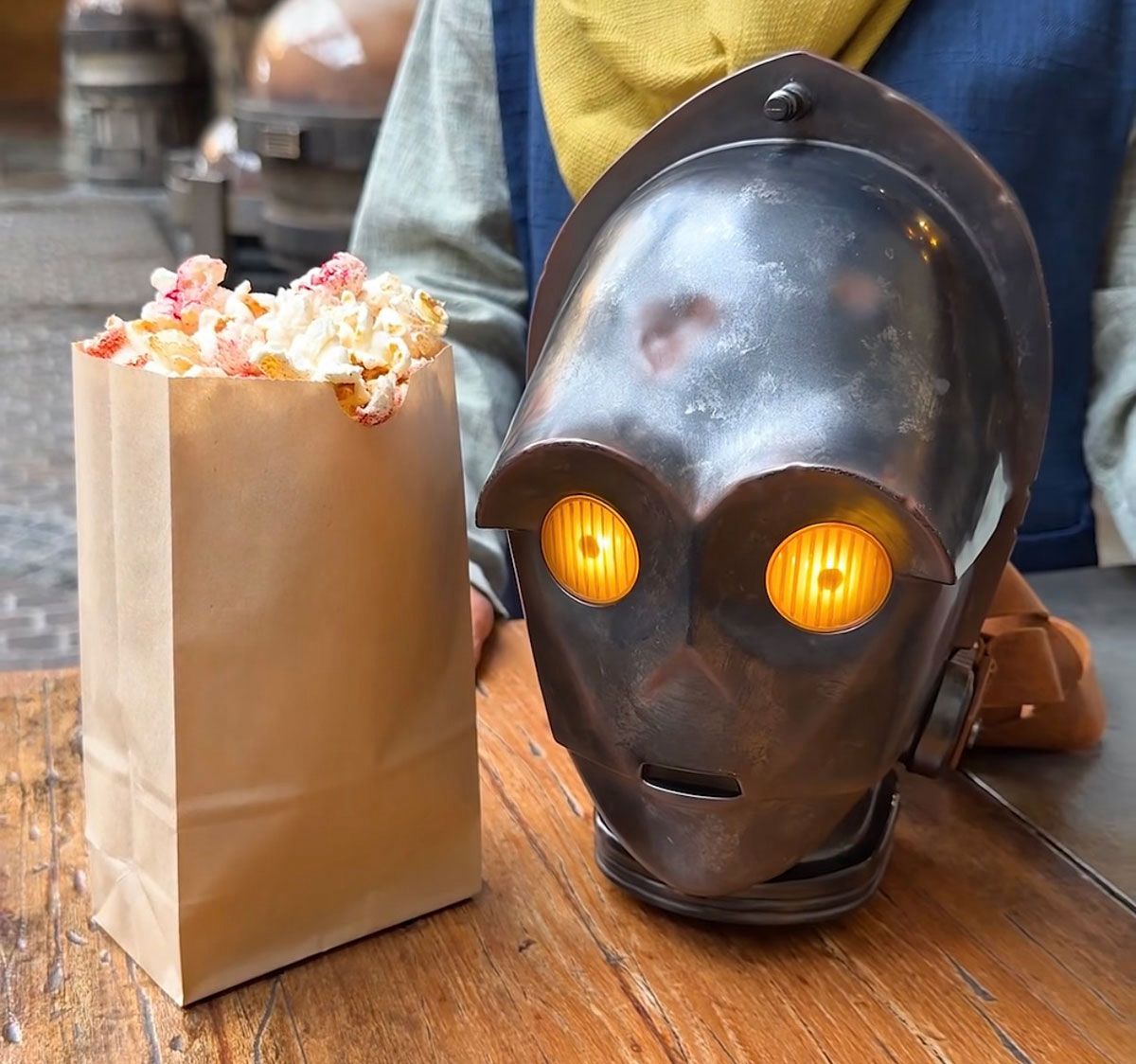 Balde de pipoca Salvaged Protocol Droid inspirado em Star Wars com olhos iluminados ao lado de pipoca temática no Disney’s Hollywood Studios durante o Star Wars Day em Orlando