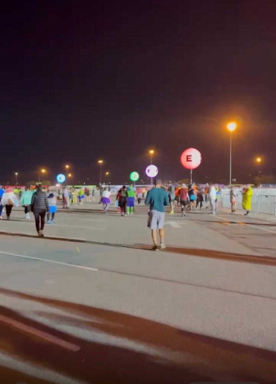 Participantes caminhando em direção aos corrais de largada de uma corrida Disney durante a madrugada, com sinalização dos grupos de saída e preparação para o início da prova runDisney