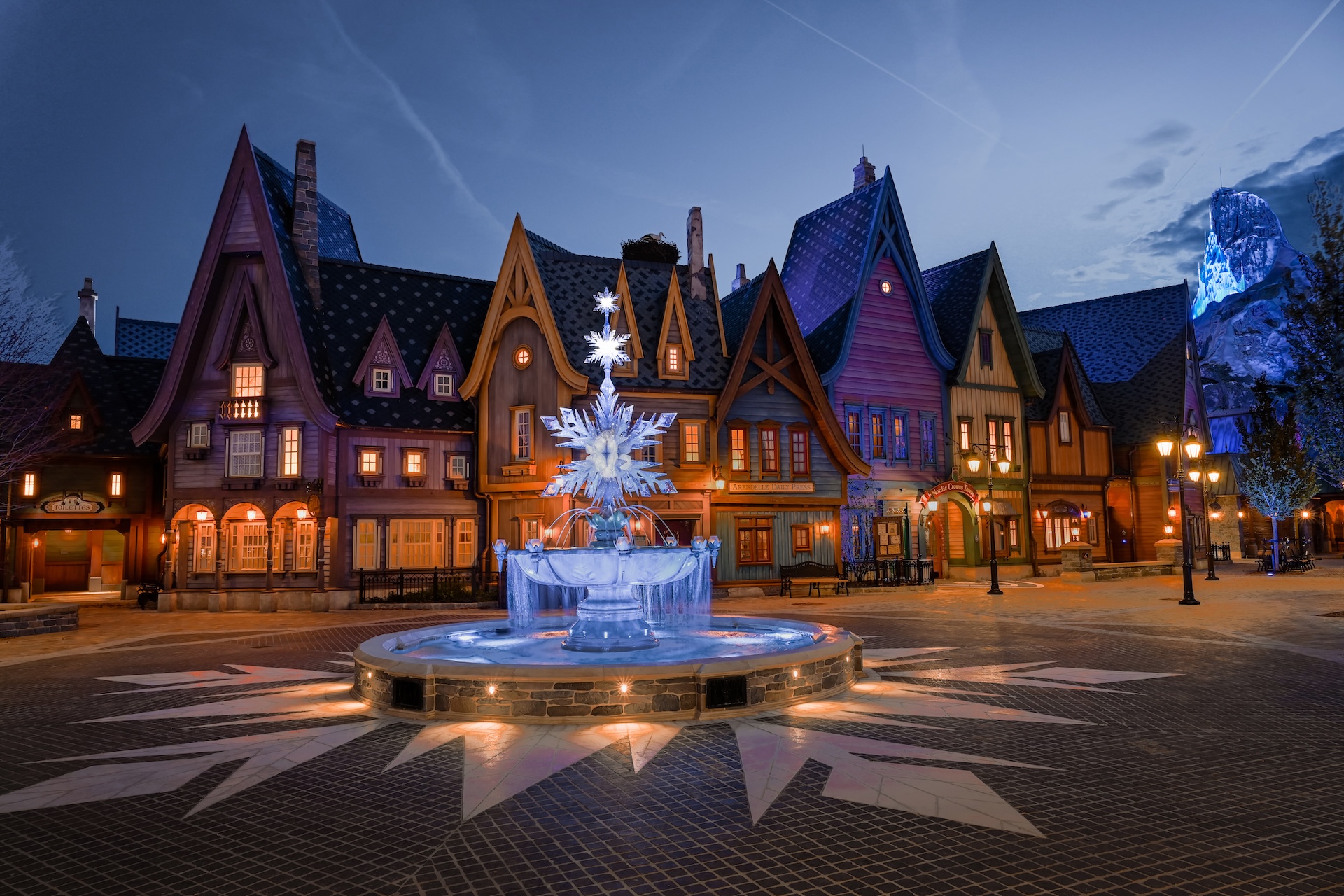 Vista noturna da área World of Frozen na Disneyland Paris, com as construções inspiradas em Arendelle, fonte iluminada e a Montanha do Norte ao fundo no Disney Adventure World