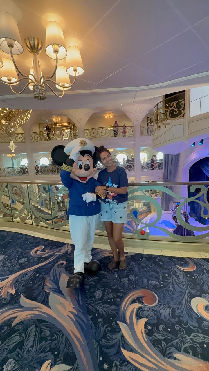 Beatriz da Talla Turismo com Mickey Mouse vestido de capitão no lobby do navio Disney Cruise Line, com decoração elegante inspirada em contos de fadas.