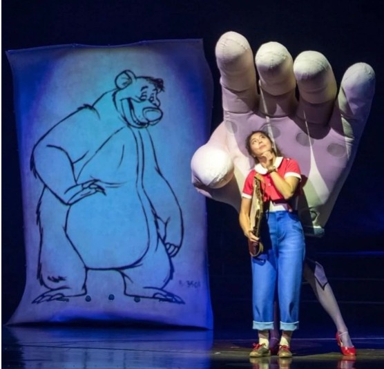 Cena do espetáculo Drawn to Life do Cirque du Soleil em Disney Springs, com personagem interagindo com uma mão gigante inspirada no processo de animação da Disney durante uma apresentação exclusiva em Orlando