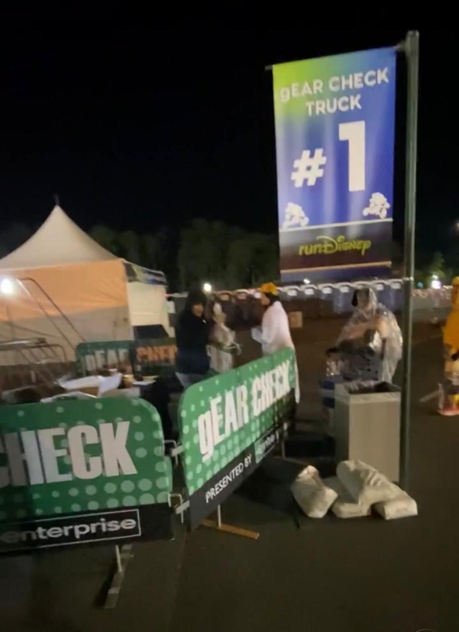 Área de gEAR Check de uma corrida runDisney durante a madrugada, com sinalização para entrega de pertences e estrutura de guarda volumes disponível para os corredores antes da largada