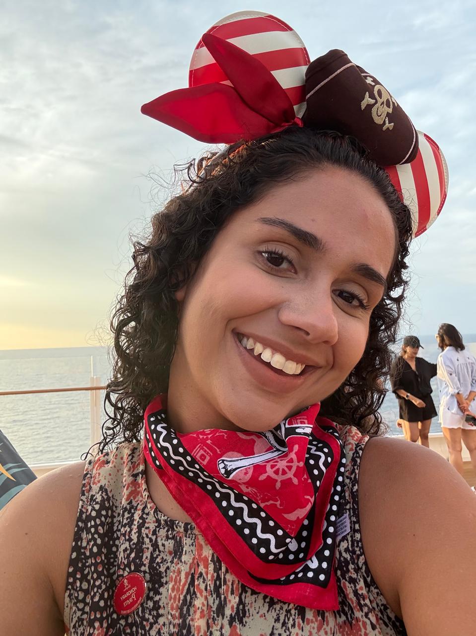 Beatriz da Talla Turismo sorrindo durante a Pirate Night no cruzeiro Disney, usando acessório temático de pirata, com o mar ao fundo ao pôr do sol.